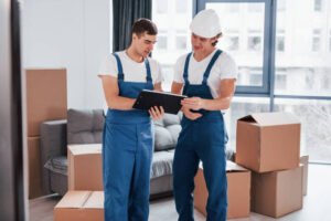 Holds notepad with document. Two young movers in blue uniform working indoors in the room.